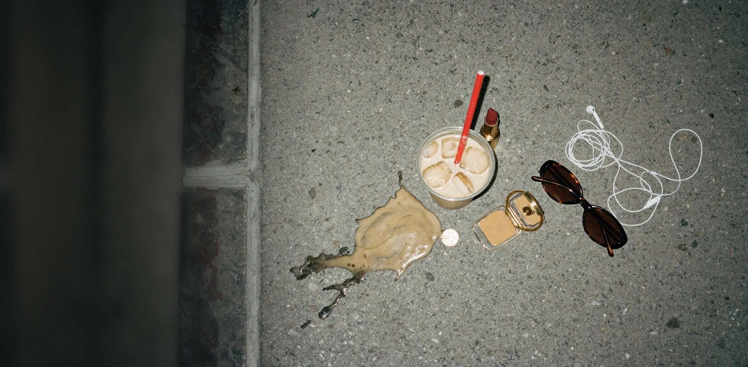 An iced coffee with a red straw spilled on a city sidewalk at night, next to a lipstick, a compact, sunglasses and earbuds.